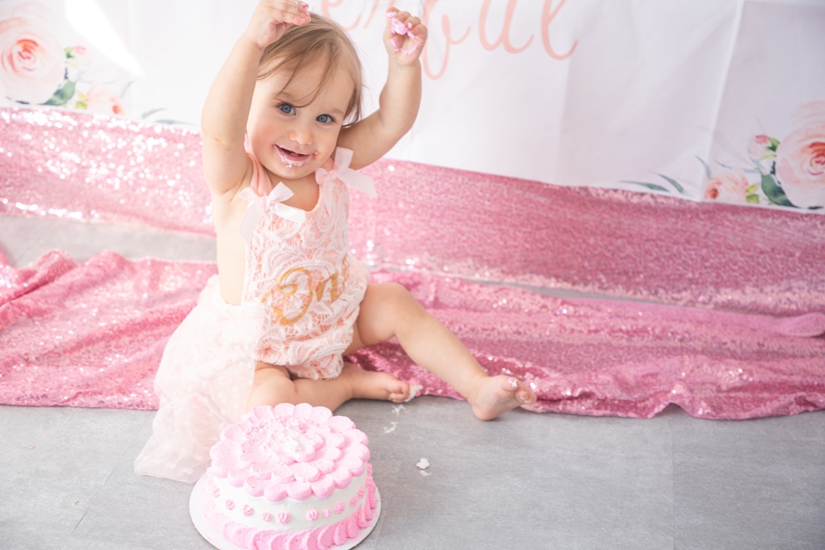 Birthday girl raising her hand with frosting on her face at her cake smash