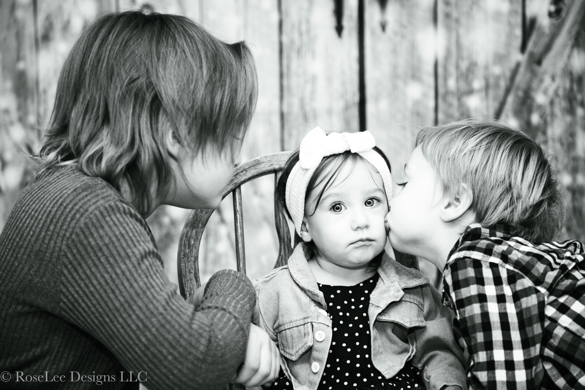 Mother and older sibling kissing a baby in black and white