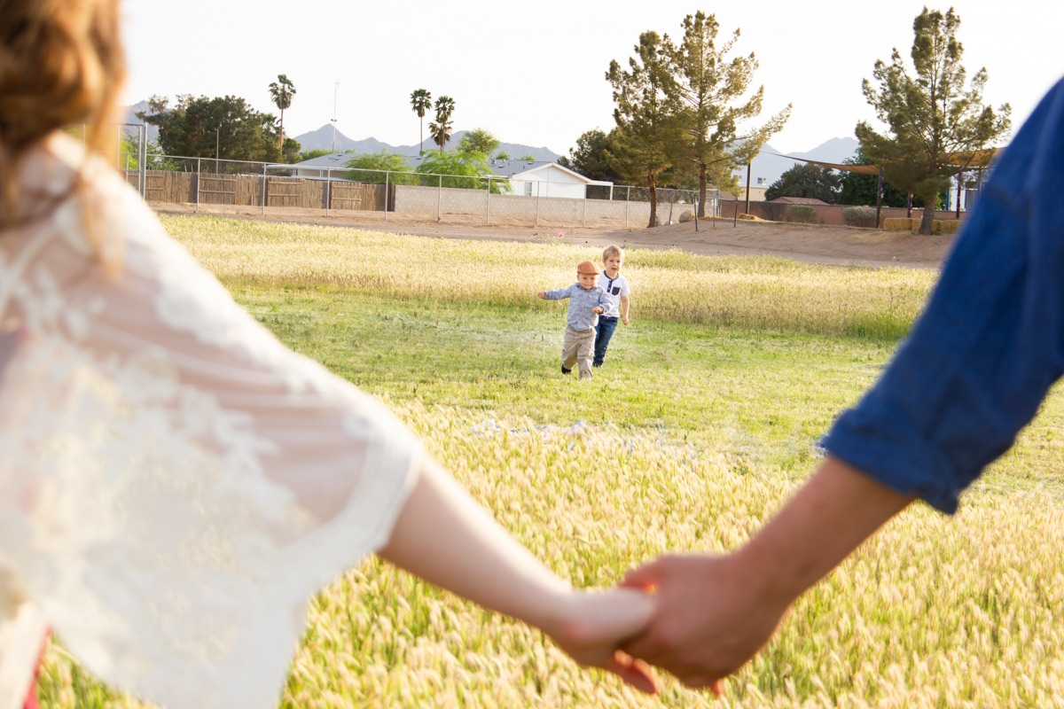 Parents holding hands in the foreground while their little boy runs through the grass