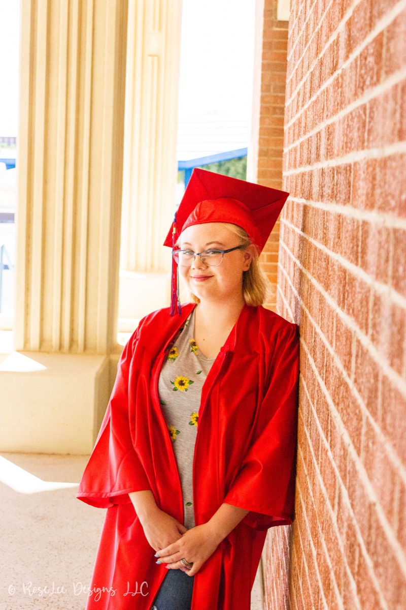 Graduate in a red cap and gown beside a brick wall